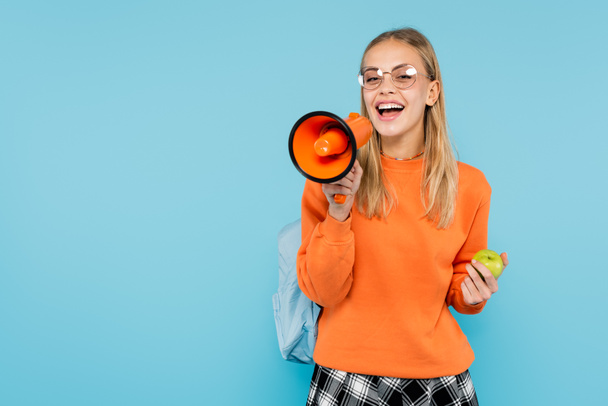 Blonde Studentin mit Brille schreit in Lautsprecher und hält Apfel isoliert auf blauem Grund  - Foto, Bild