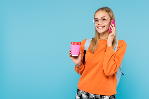 Estudiante sonriente en anteojos hablando por celular y sosteniendo café para ir aislado en azul  - Foto, imagen