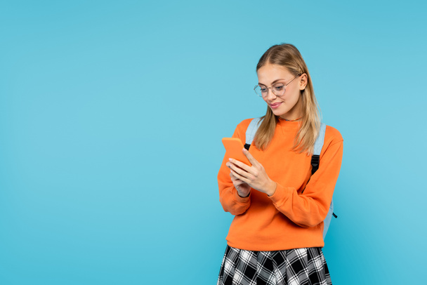 Blonde student with backpack and eyeglasses using mobile phone isolated on blue  - Foto, immagini