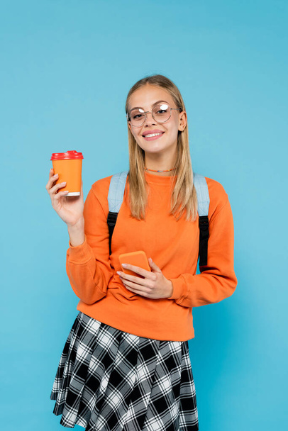 Positive student in eyeglasses holding smartphone and paper cup isolated on blue  - 写真・画像