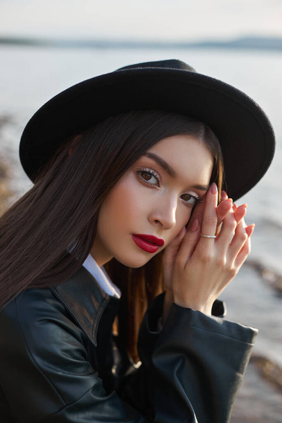 Portrait young woman at sunset. Fashion woman in black round hat and leather raincoat near lake pond. Beautiful makeup and bright red lipstick on lips - Фото, изображение