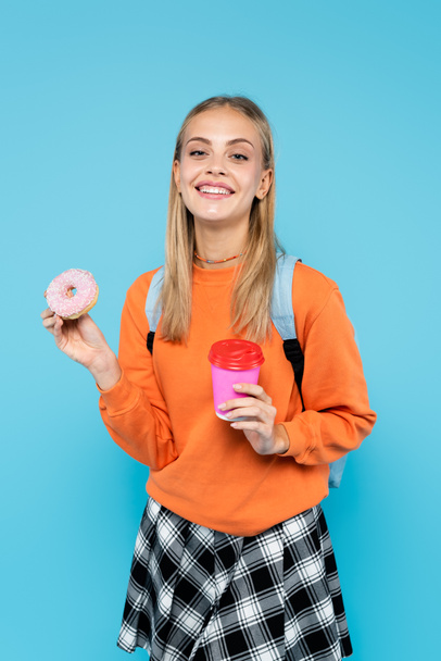 Cheerful student with backpack holding paper cup and donut isolated on blue  - Фото, изображение