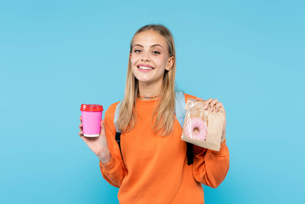 Cheerful student holding donut in package and takeaway drink isolated on blue  - Foto, Bild