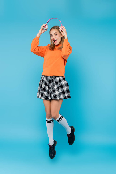 Cheerful student holding headphones while jumping on blue background  - Foto, afbeelding