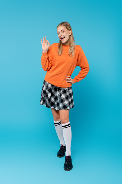 Full length of smiling student in skirt showing okay at camera on blue background  - Fotó, kép