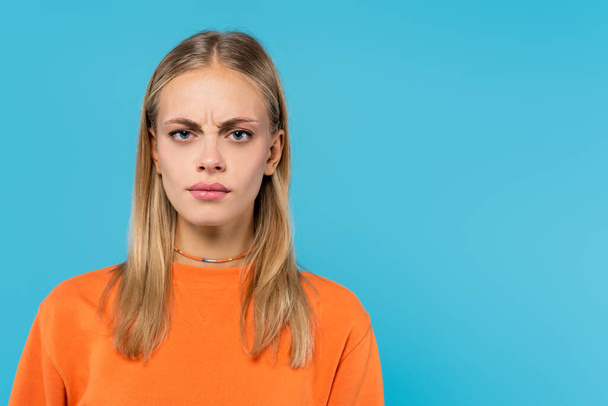 Offended blonde woman in orange sweatshirt looking at camera isolated on blue  - Fotó, kép