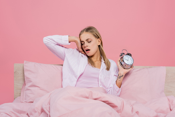Szőke nő pizsamában tartja ébresztőórát és ásít az ágyon elszigetelt rózsaszín  - Fotó, kép