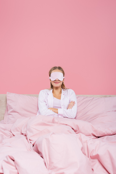 Mulher sorridente em máscara de sono sentado com os braços cruzados na cama isolado em rosa  - Foto, Imagem