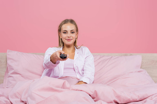 glimlachende vrouw in pyjama houden afstandsbediening op bed geïsoleerd op roze  - Foto, afbeelding