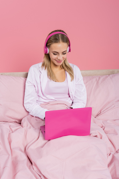 Positive Frau in Kopfhörer und Schlafanzug mit Laptop auf dem Bett isoliert auf rosa  - Foto, Bild