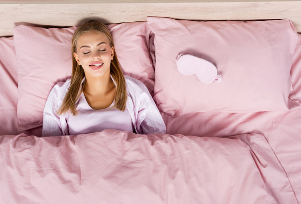 Vue du dessus de la femme blonde qui sort la langue près du masque de sommeil sur le lit  - Photo, image