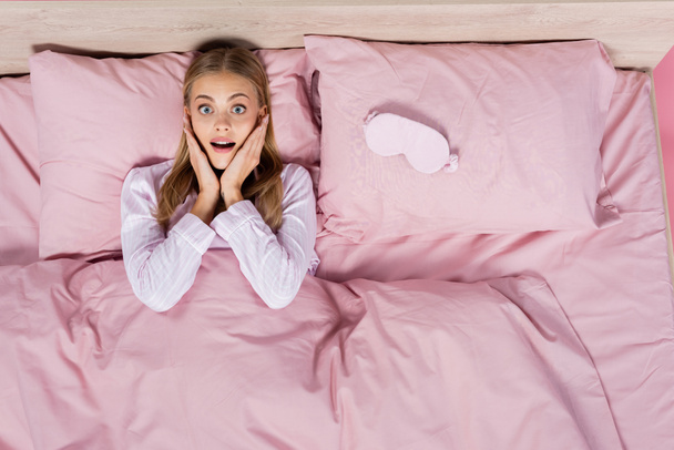 Horní pohled na vzrušenou ženu v pyžamu při pohledu na kameru na posteli izolované na růžové  - Fotografie, Obrázek