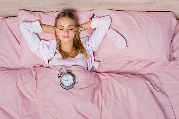Top view of woman lying near alarm clock and sleep mask on bed  - Photo, Image