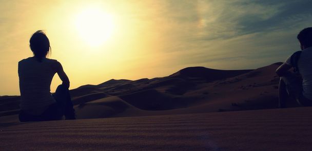 Пара молодых авантюристов, сидящих над песчаной дюной и наслаждающихся удивительным закатом над красными дюнами моря Эрг Чебби, Марокко
. - Фото, изображение