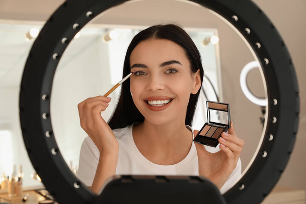 Beautiful young woman applying eye shadow with brush indoors, view through ring lamp - Fotoğraf, Görsel