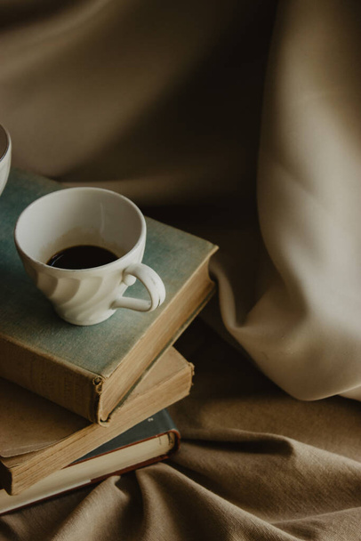 A vertical shot of a coffee cup on piled old books and beige fabric surface - 写真・画像