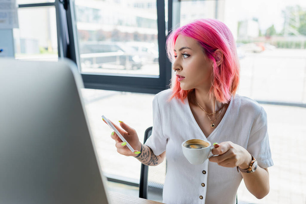 mladý piercing manažer s růžovými vlasy drží šálek kávy a smartphone v kanceláři - Fotografie, Obrázek