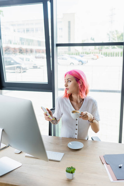 doorboorde manager met roze haar holding kopje koffie en smartphone in het kantoor - Foto, afbeelding