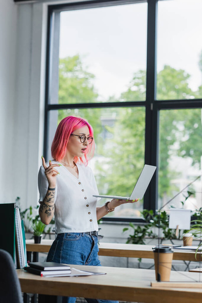 veselá mladá podnikatelka s růžovými vlasy pomocí notebooku během videohovoru v kanceláři - Fotografie, Obrázek