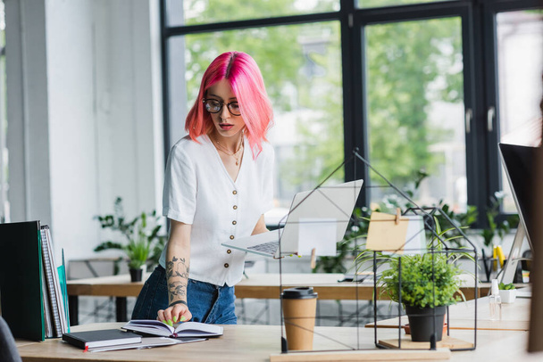 young businesswoman with pink hair holding laptop and looking at notebook  - Фото, зображення