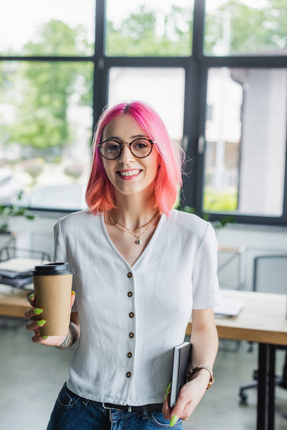 cheerful and pierced businesswoman with pink hair holding paper cup and notebook - Photo, Image