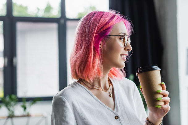 side view of pierced businesswoman with pink hair holding paper cup and smiling in office - Photo, Image