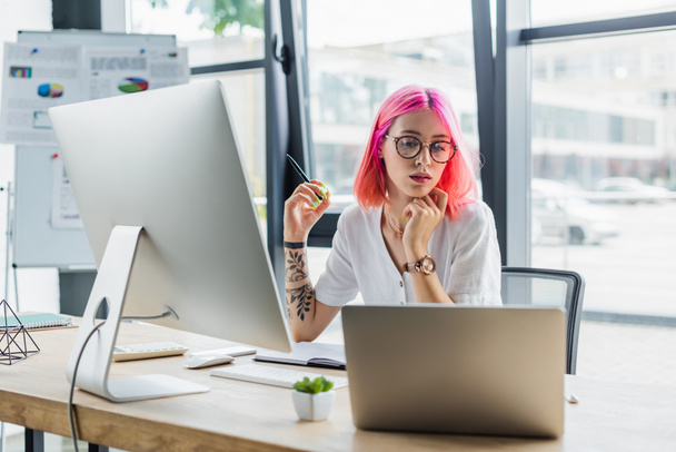 pierced businesswoman with pink hair holding pen and looking at laptop near computer monitor - Foto, afbeelding