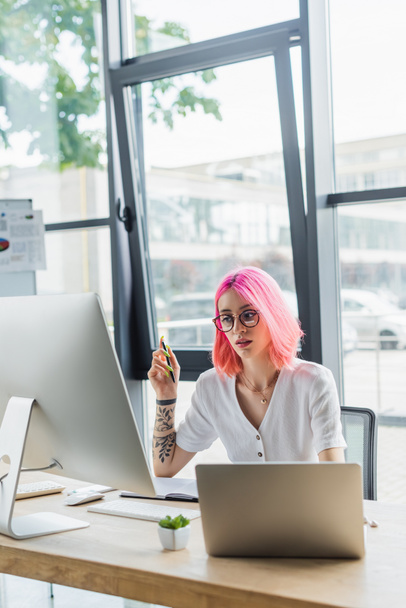 doorboorde zakenvrouw met roze haar holding pen en op zoek naar computer monitor - Foto, afbeelding