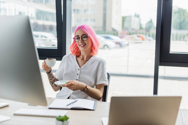 happy businesswoman with pink hair holding cup of coffee and looking at computer monitor - Photo, Image