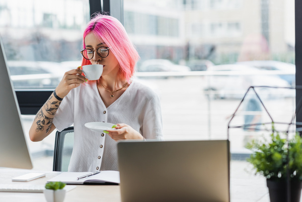 mulher de negócios tatuada com cabelo rosa bebendo café perto do monitor do computador - Foto, Imagem