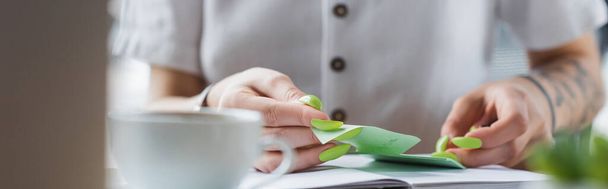 cropped view of tattooed businesswoman holding sticky note near notebook and blurred cup, banner - Фото, изображение