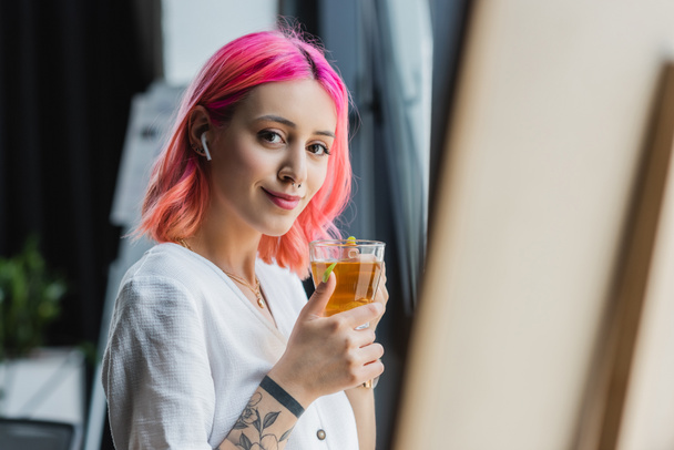 cheerful businesswoman with pink hair and earphone holding cup of tea in office - Foto, Imagen