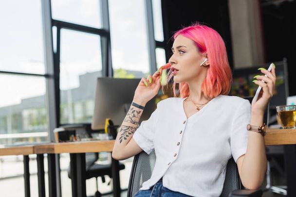 tattooed businesswoman with pink hair and earphone holding smartphone in office - Foto, Bild