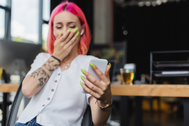 размытая деловая женщина с розовыми волосами и наушниками с помощью смартфона и прикрывая рот во время смеха в офисе - Фото, изображение