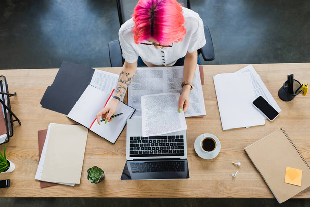 вид на деловую женщину с розовыми волосами, держащую ручку рядом с документами и гаджетами на столе - Фото, изображение