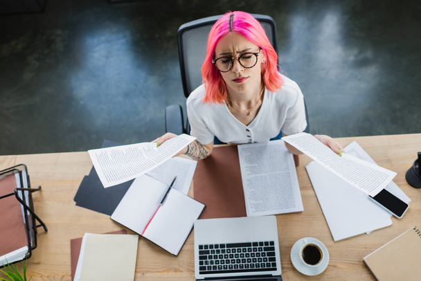 top view of tired businesswoman with pink hair holding documents near gadgets on desk - Photo, Image
