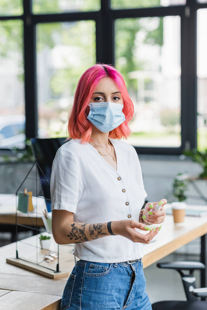 mujer de negocios tatuada con desinfectante de pulverización de cabello rosa en la mano en la oficina - Foto, Imagen