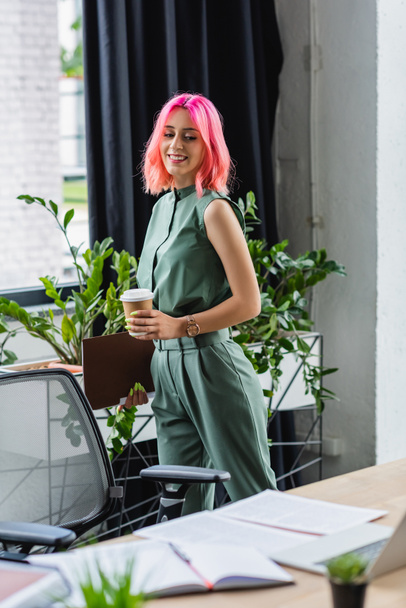 cheerful manager with pink hair and piercing holding paper cup and folder while looking at desk - Foto, Imagen