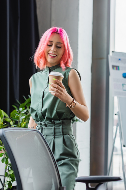 cheerful manager with pink hair and piercing holding paper cup while looking at office chair - Fotó, kép