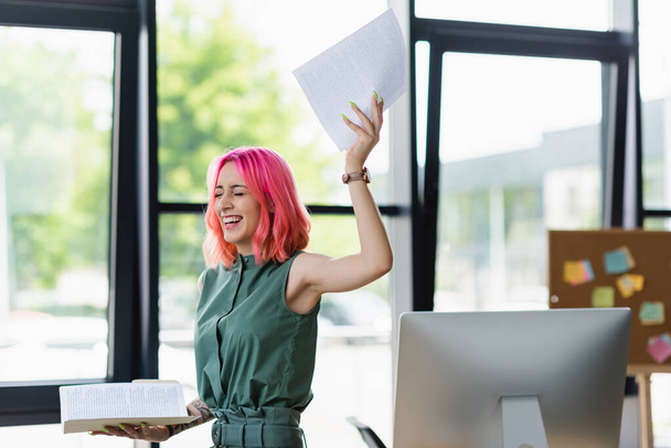 cheerful businesswoman with pink hair and piercing holding folder with documents in office - Zdjęcie, obraz