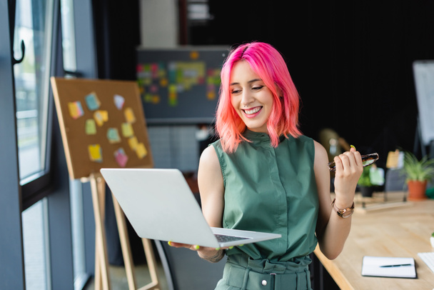 joyful businesswoman with pink hair holding laptop and eyeglasses in office - Photo, Image
