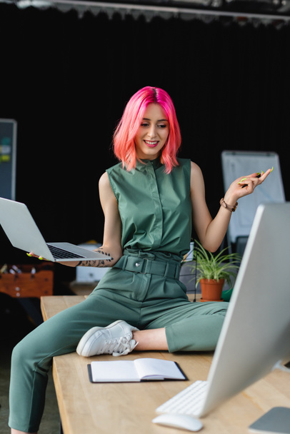 örömteli üzletasszony rózsaszín haj gazdaság laptop és nézi monitor az irodában - Fotó, kép