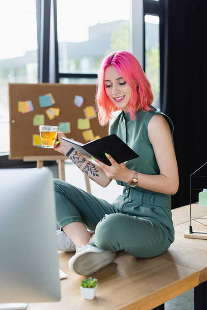 happy businesswoman with pink hair holding cup of tea and looking at notebook while sitting on desk - Foto, Bild