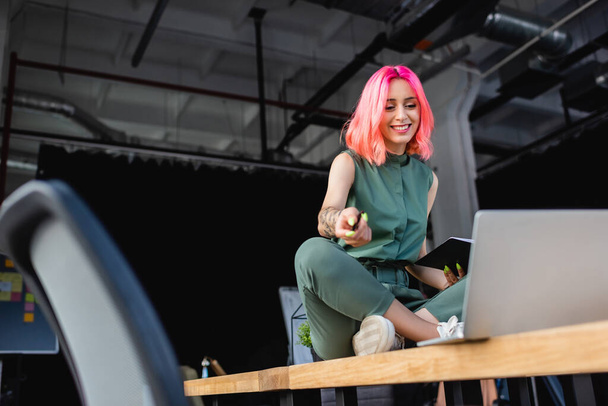 szczęśliwy businesswoman z różowymi włosami trzymając długopis i notatnik podczas siedzenia na biurku i patrząc na laptopa - Zdjęcie, obraz