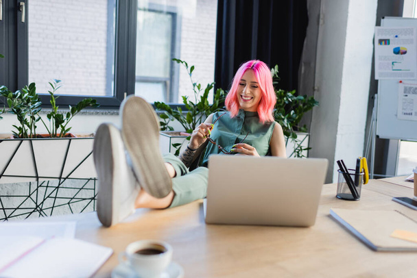 mulher de negócios feliz com cabelo rosa segurando óculos enquanto olha para laptop  - Foto, Imagem