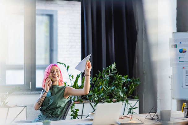positive businesswoman with pink hair talking on smartphone while throwing paper plane - Fotó, kép