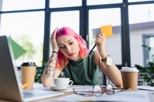 zmęczony menedżer z różowymi włosami trzymając długopis w pobliżu papierowych filiżanek z kawą w biurze - Zdjęcie, obraz