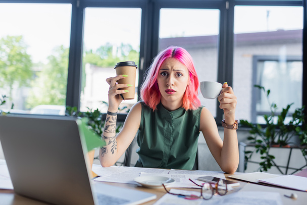 shocked manager with pink hair holding coffee near devices in office - Foto, imagen