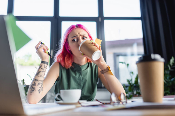 femme d'affaires choquée avec les cheveux roses boire du café près d'un ordinateur portable au bureau - Photo, image