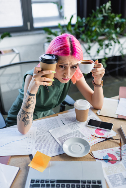 high angle view of stressed businesswoman with pink hair holding coffee and looking at laptop  - Foto, afbeelding